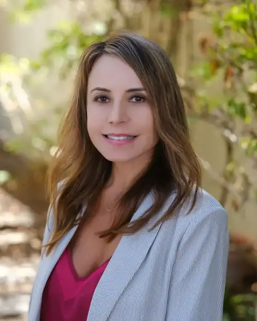 Professional woman outdoors, wearing a light blazer and pink top, smiling at the camera amid greenery.