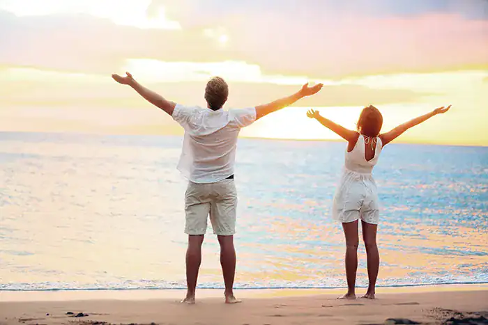 Couple standing on a sandy beach at sunset with arms raised in celebration, facing the water.