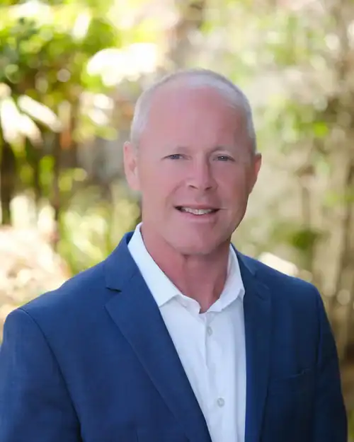 Portrait of a smiling man in a blue suit jacket and white shirt outdoors with greenery in the background, suggesting a professional headshot.