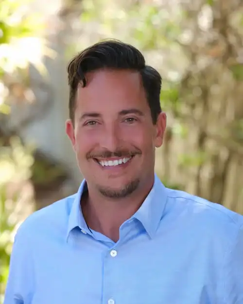 Smiling man in a light blue button-down shirt outdoors, with greenery in the background.