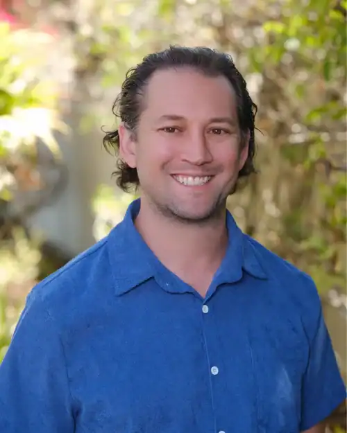 Smiling man with dark, wavy hair wearing a blue button-down shirt, standing outdoors with greenery in the background.