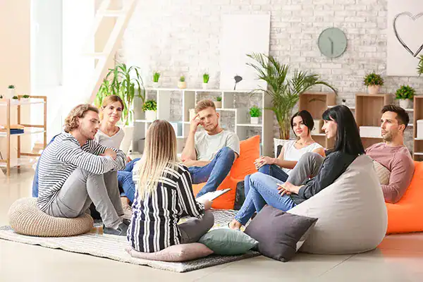 Eight people sitting in a circle on floor cushions in a bright living room, chatting in a casual group discussion.