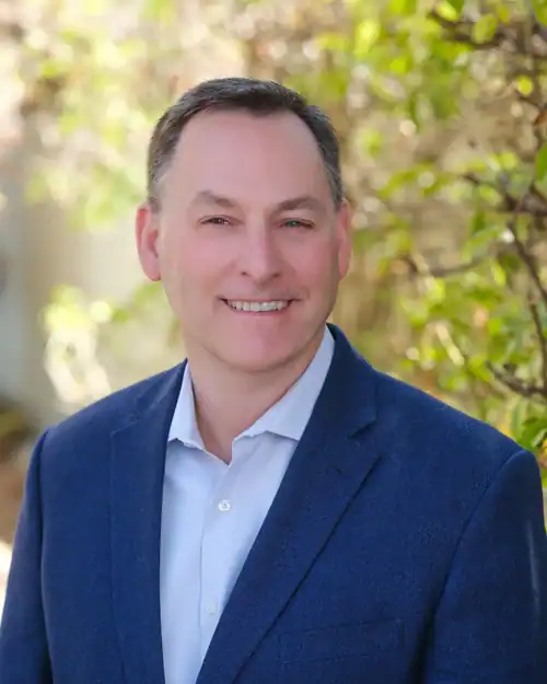 Professional headshot of a smiling man in a blue blazer outdoors, sunlit greenery in the background.