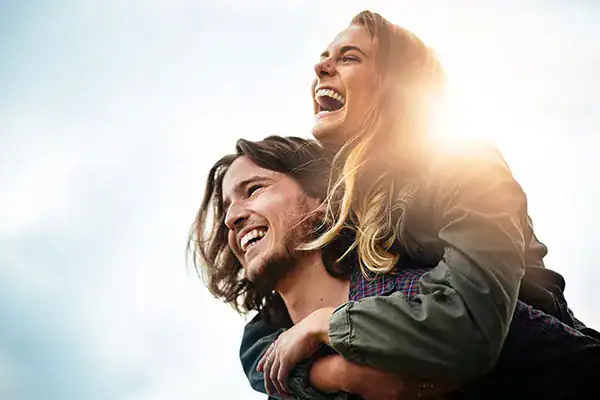 Couple smiling and laughing as one person carries the other on their back outdoors with sunlight in the background.
