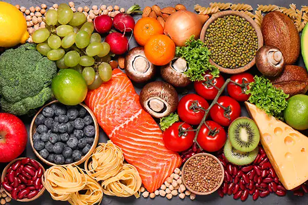 Colorful flat lay of assorted healthy foods: salmon fillet center, fruits, vegetables, grains, beans, and cheese arranged on a dark background.
