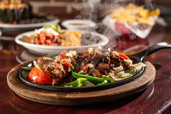 Sizzling steak fajitas on a hot cast-iron skillet with bell peppers and onions, steam rising, in a restaurant setting