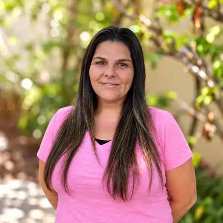 Cassandra P - Right Path Recovery San Diego - Right Path Recovery San Diego Woman with long dark hair wearing a pink T-shirt, standing outdoors with green plants behind her, smiling.