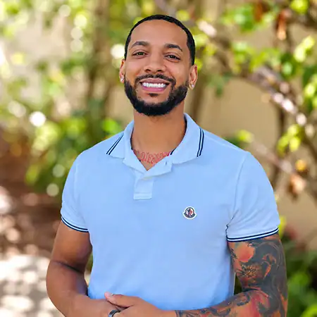 Devon D - Right Path Recovery San Diego - Right Path Recovery San Diego Smiling man with a beard and tattoos on his left arm, wearing a light blue polo shirt outdoors among greenery.