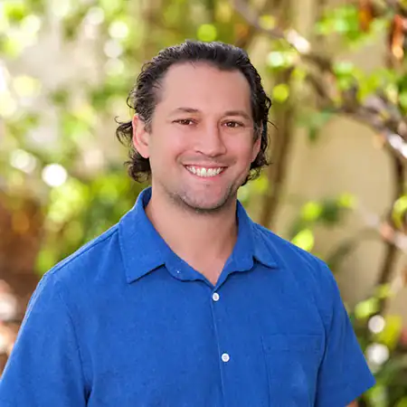 Dr Troy Kurz copy - Right Path Recovery San Diego - Right Path Recovery San Diego Smiling man in a blue button-down shirt standing outdoors with greenery in the background.