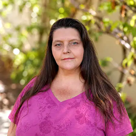 Jaimie M - Right Path Recovery San Diego - Right Path Recovery San Diego Portrait of a woman with long dark hair standing outdoors in a garden, wearing a pink patterned T-shirt, with a neutral expression in the camera.