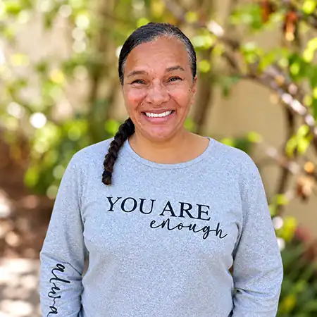 Jamillah - Right Path Recovery San Diego - Right Path Recovery San Diego Smiling woman with a braided ponytail wearing a light gray shirt that reads 'YOU ARE enough' outdoors.