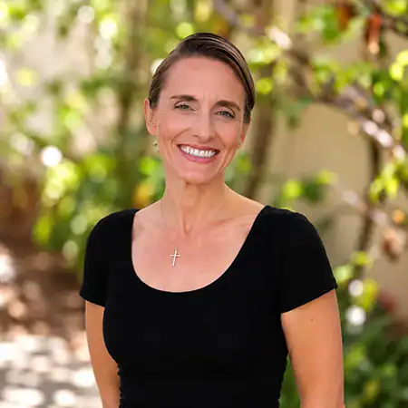 Janelle Neeley - Right Path Recovery San Diego - Right Path Recovery San Diego Smiling woman outdoors wearing a black short-sleeve top and a cross necklace, with greenery in the blurred background.