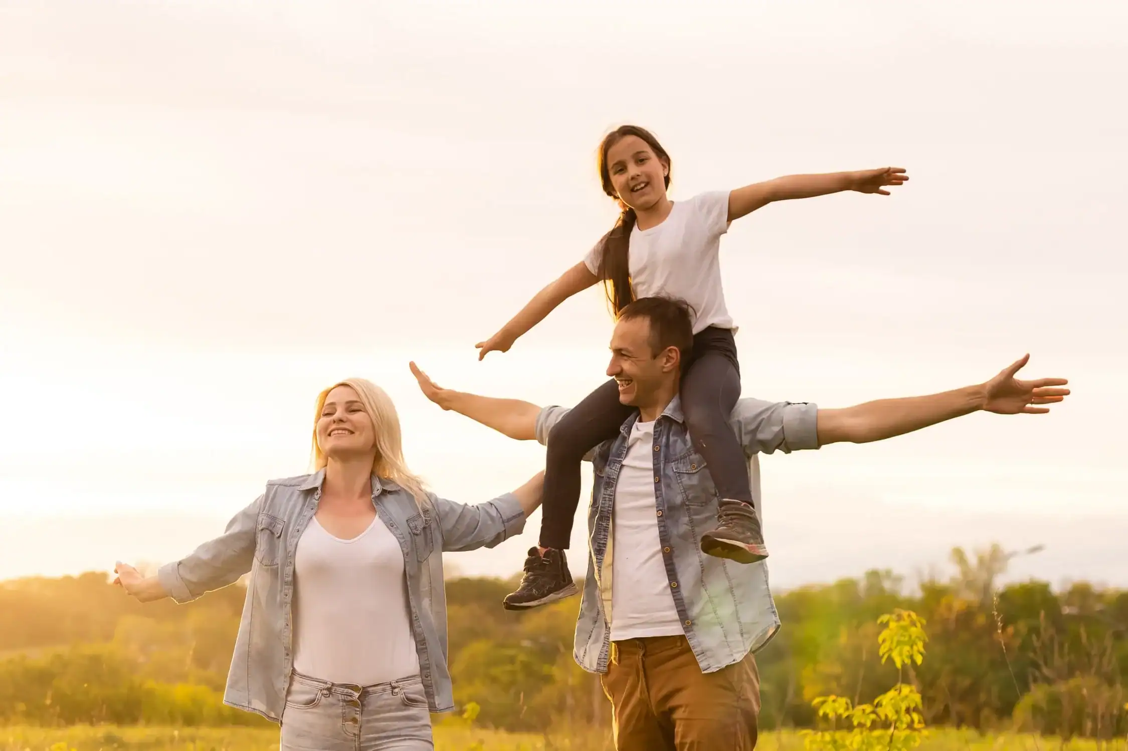 Family of three outdoors at sunset: dad carrying daughter on his shoulders while mom walks beside with arms outstretched, all smiling in a field at dusk.