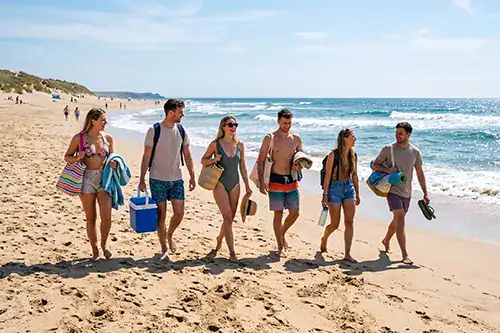 Group of friends walking along a sunny beach, carrying towels, bags, and a cooler toward the water.