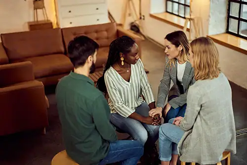 A diverse group of four adults seated in a circle, one woman shaking hands with another as they talk in a casual meeting space.
