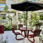 Right_Path_RecoveryRio_IOP41 - Right Path Recovery San Diego - Right Path Recovery San Diego Outdoor courtyard seating with a round red table and four matching chairs under a large black umbrella amid greenery in a modern building complex.