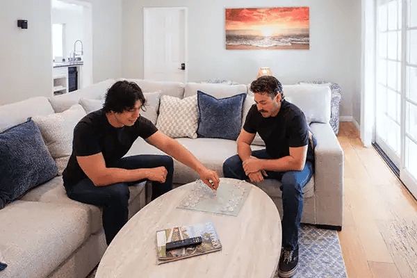 Two men sit on a light gray sectional in a bright living room, playing a board game at a wooden coffee table.